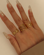 Mano con anillos dorados de bandas entrecruzadas y flores, sobre fondo beige.