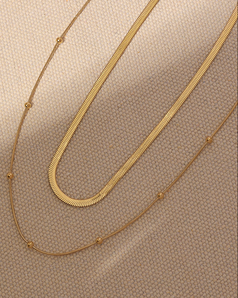 Detalle de dos cadenas doradas, una plana y otra fina con pequeñas esferas, extendidas sobre tela beige.