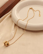 Collar dorado con colgante geométrico de bloques y detalles oscuros, junto a un plato blanco sobre tela clara.
