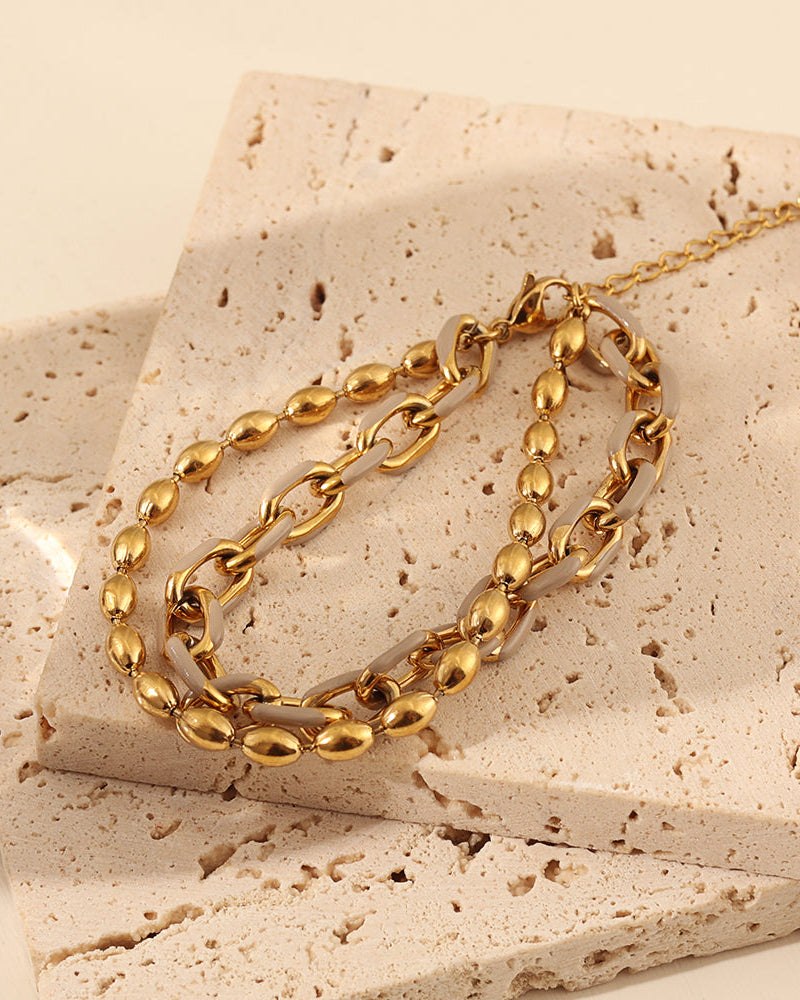 Pulsera dorada de doble cadena con eslabones ovalados y detalles oscuros sobre una losa de piedra clara.