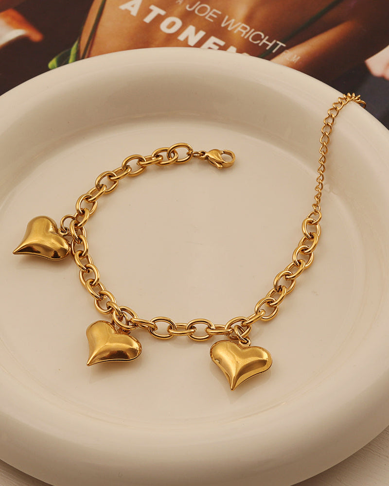 Pulsera dorada de cadena con tres corazones lisos colgantes, sobre un plato blanco.