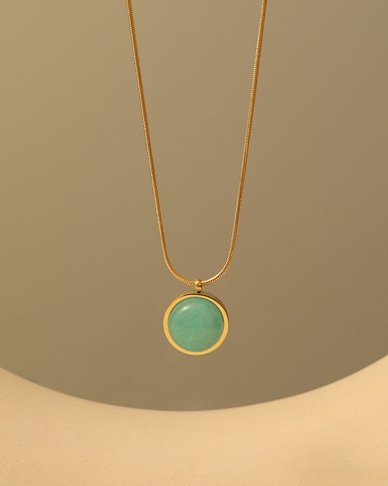 Collar fino dorado con colgante circular verde de borde dorado, sobre un expositor beige.