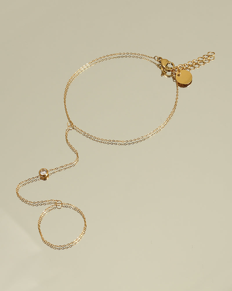 Pulsera fina dorada con cadena hacia un aro para el dedo y pequeñas cuentas brillantes, extendida sobre fondo claro.