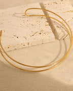 Collar rígido dorado de dos aros finos abiertos, sobre tela beige y losas de piedra clara.