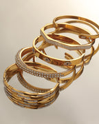 Conjunto de pulseras rígidas doradas apiladas, con diseños lisos, cruzados y cubiertos de pequeñas piedras claras.