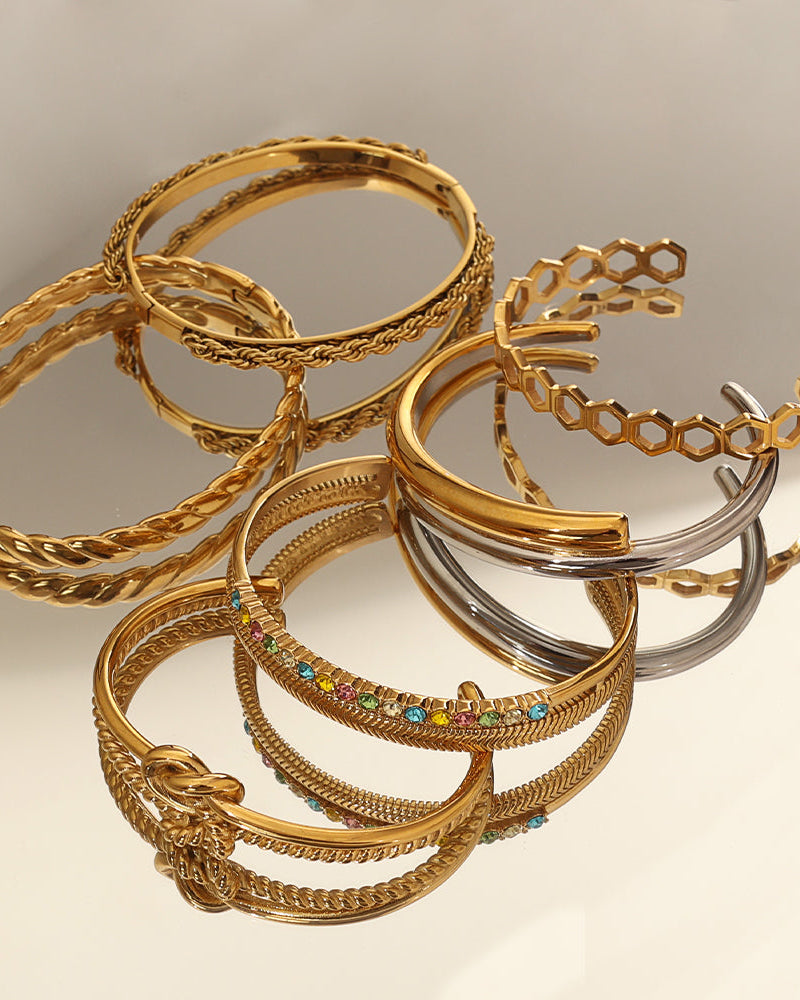 Varias pulseras rígidas doradas, una con nudo y otras con piedras y detalles oscuros, sobre superficie reflectante.