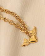 Detalle de collar dorado de eslabones ovalados y colgante de corazón con alas extendidas, sobre fondo claro.