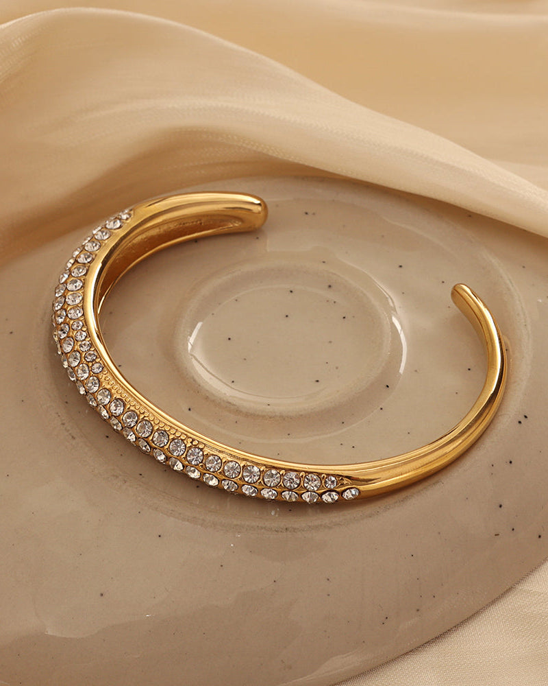 Pulsera rígida abierta dorada con varias hileras de pequeñas piedras claras, sobre plato de cerámica beige.