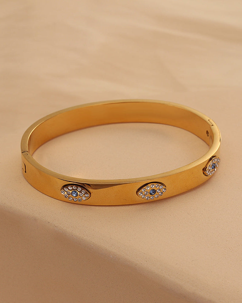 Pulsera rígida ancha dorada con motivos ovalados de pequeñas piedras claras, sobre tela beige.