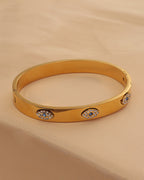 Pulsera rígida ancha dorada con motivos ovalados de pequeñas piedras claras, sobre tela beige.