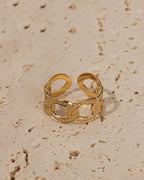 Anillo abierto dorado de grandes eslabones ovalados con pequeñas piedras claras, sobre piedra beige.
