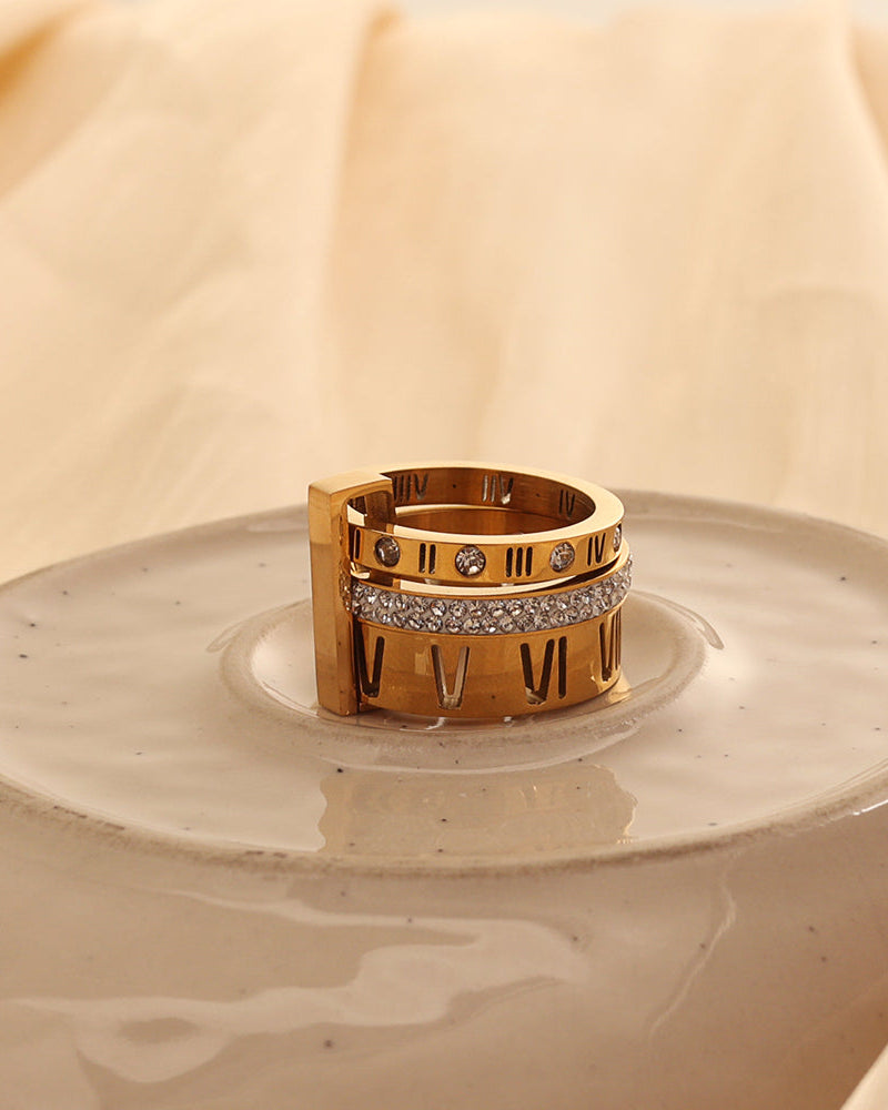 Anillo ancho dorado con números romanos oscuros y franja de pequeñas piedras claras, sobre plato de cerámica.