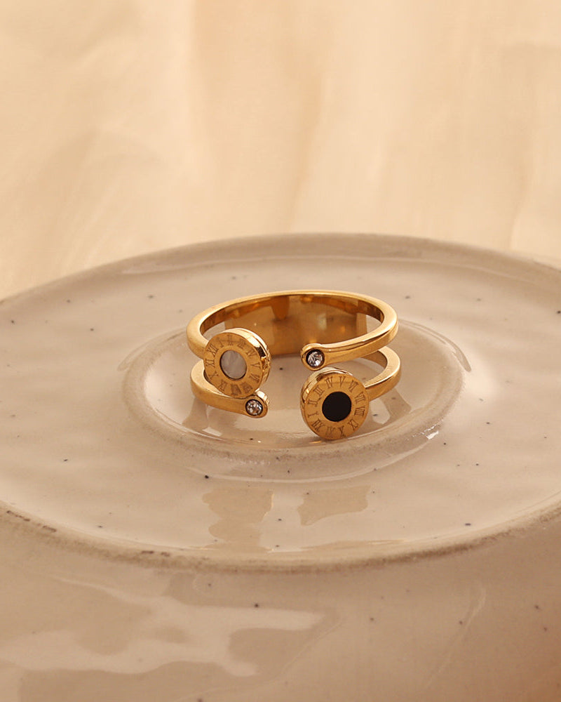 Anillo abierto dorado con dos discos, uno de centro negro y otro claro, y pequeñas piedras en los extremos, sobre plato de cerámica.