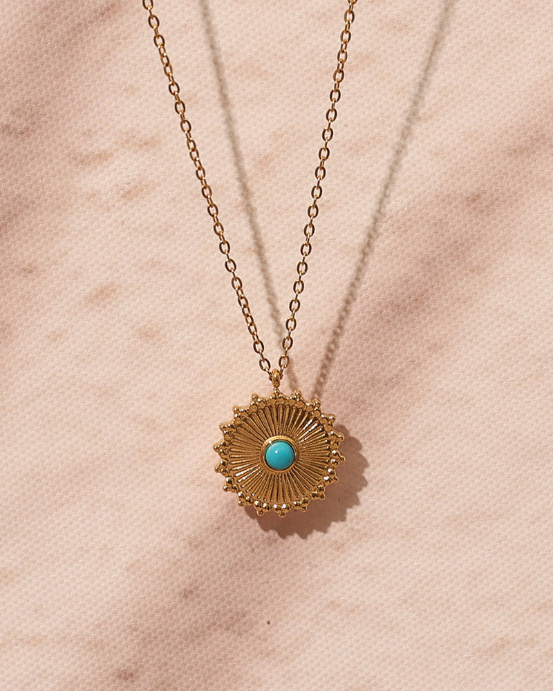 Collar fino dorado con medallón de rayos y centro azul turquesa sobre superficie rosada.