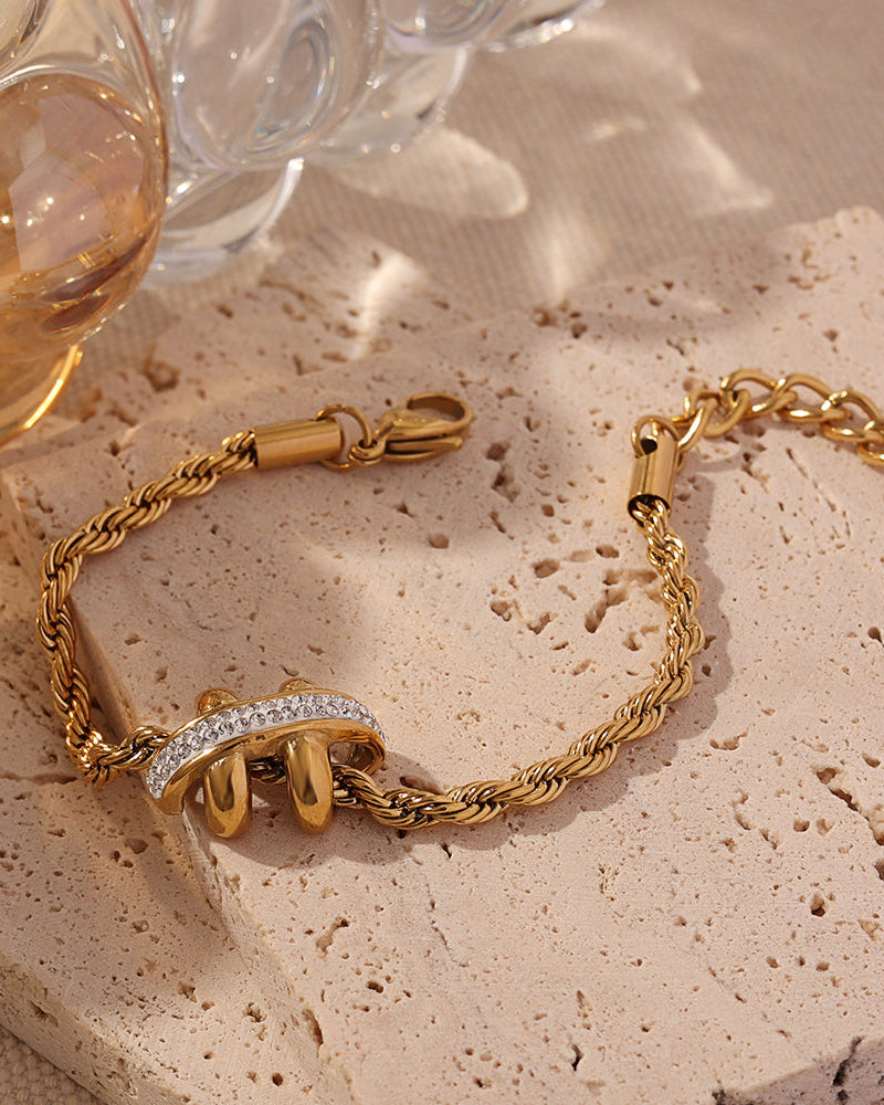 Pulsera dorada de cadena torcida con adorno central de lazos y pequeñas piedras claras sobre piedra beige, junto a un vaso.
