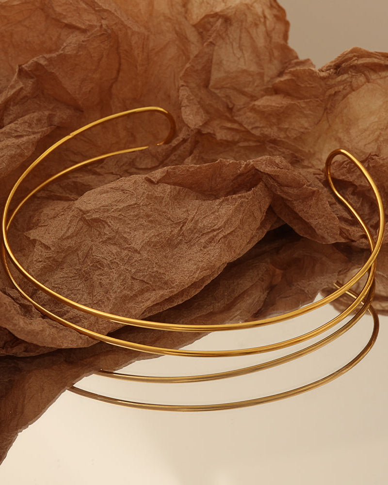 Collar rígido abierto dorado de dos aros finos, sobre papel marrón arrugado.