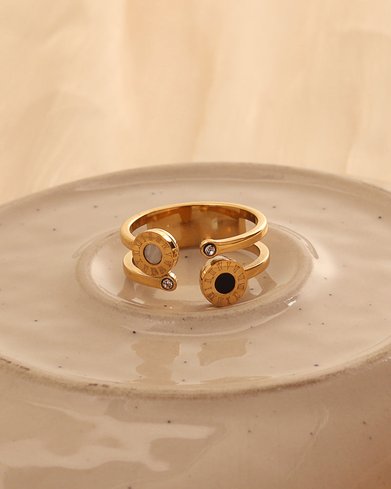 Anillo abierto dorado con discos de centro negro y claro y pequeñas piedras en los extremos, sobre plato de cerámica.