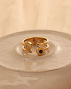 Anillo abierto dorado con discos de centro negro y claro y pequeñas piedras en los extremos, sobre plato de cerámica.