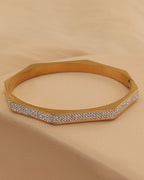 Pulsera rígida dorada de forma poligonal con varias hileras de pequeñas piedras claras en el frente, sobre tela beige.