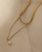 Collar de dos cadenas doradas, una plana y otra fina con perla blanca colgante, sobre tela translúcida clara.