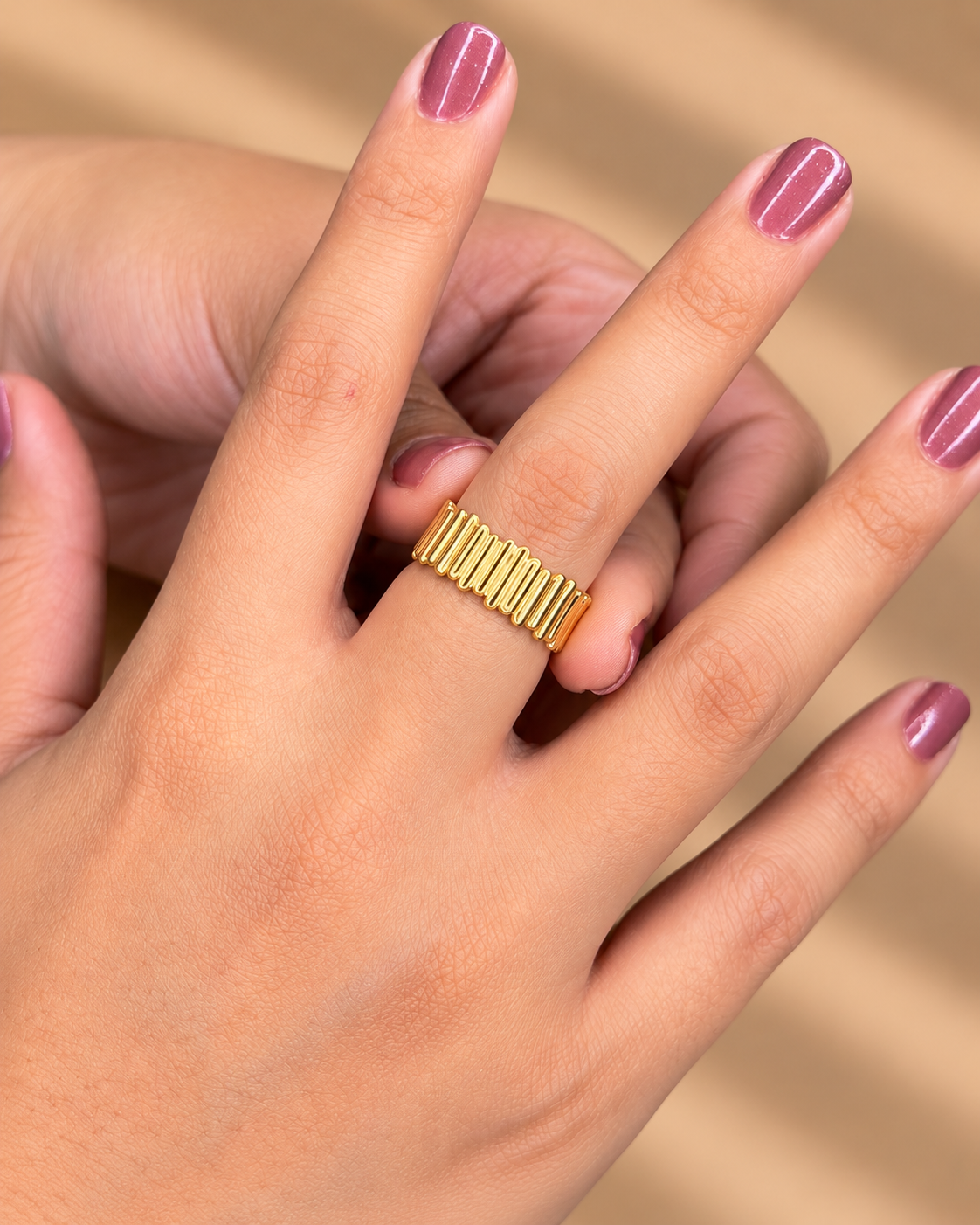 Anillo ancho dorado con estrías verticales, puesto en una mano con uñas de tono morado.