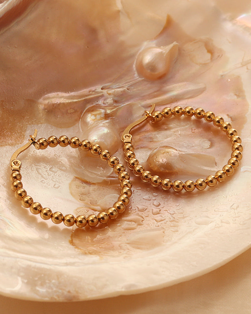 Par de pendientes dorados de aro de pequeñas esferas, sobre una concha de tonos crema.