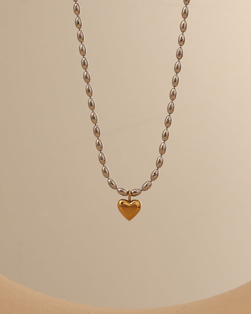Collar de cuentas ovaladas de tono plateado con pequeño corazón dorado colgante, sobre fondo beige.