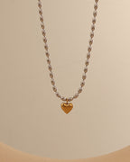 Collar de cuentas ovaladas de tono plateado con pequeño corazón dorado colgante, sobre fondo beige.