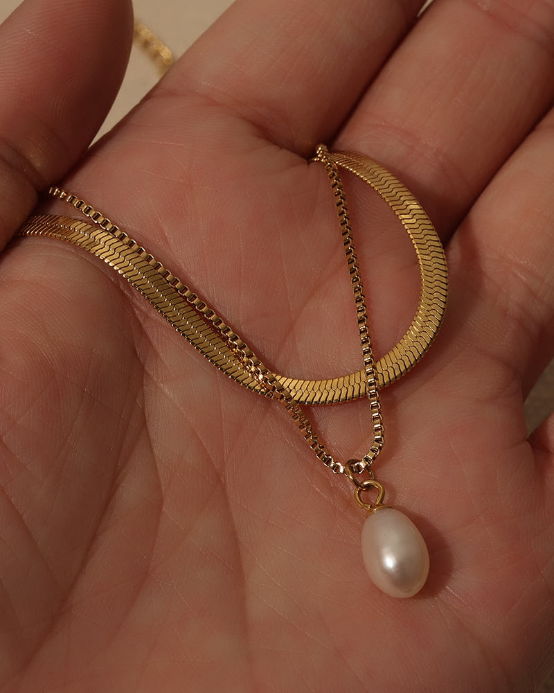 Dos cadenas doradas con colgante de perla blanca ovalada sostenidas en la palma de una mano.