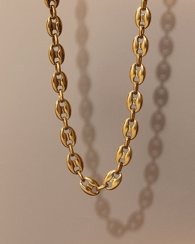 Collar dorado de eslabones ovalados gruesos, suspendido sobre fondo beige con sombra lateral.