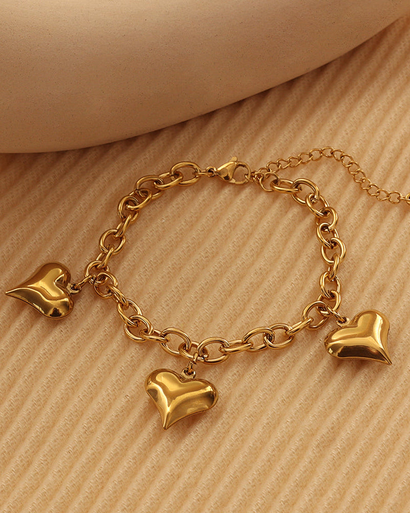 Pulsera dorada de eslabones ovalados con tres corazones colgantes y cadena de extensión, sobre tela beige.