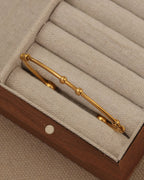 Pulsera rígida dorada de aro fino con pequeñas esferas, en un compartimento de joyero de tela beige.