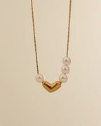 Collar fino dorado con corazón central liso y pequeñas perlas blancas a ambos lados, suspendido sobre fondo crema.