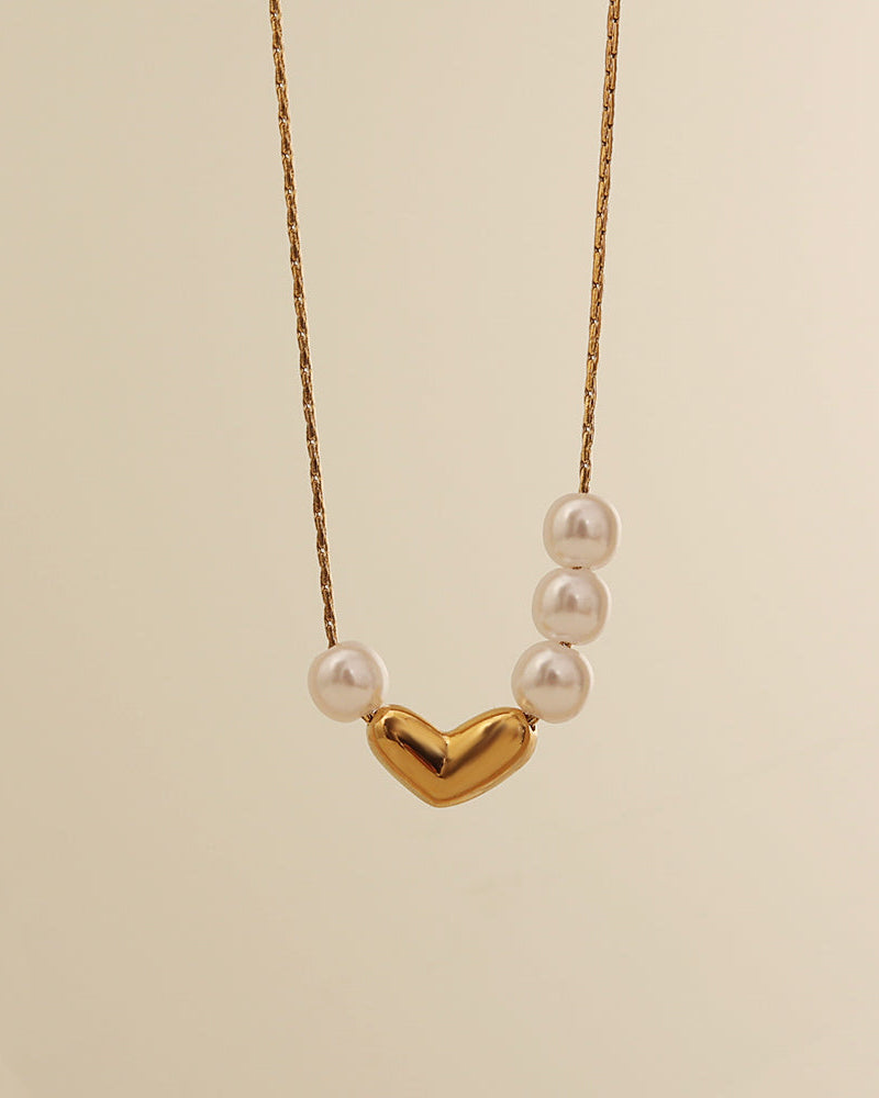 Collar fino dorado con corazón central liso y pequeñas perlas blancas a ambos lados, suspendido sobre fondo crema.
