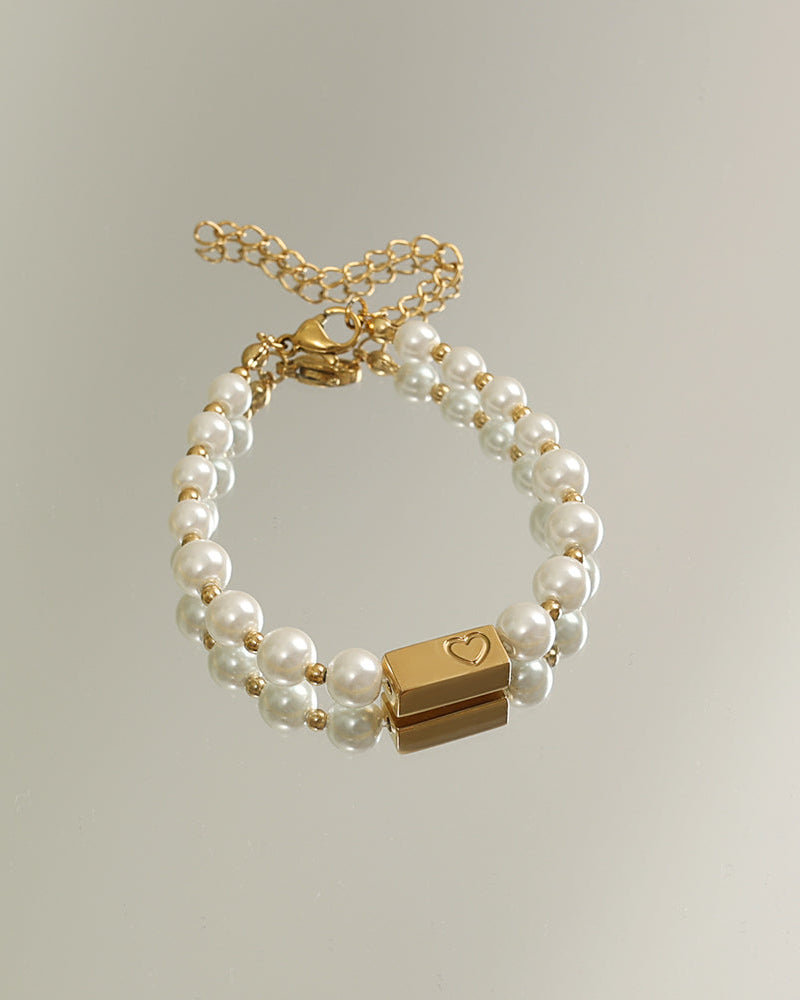 Pulsera de dos hileras de perlas blancas con adorno rectangular dorado y cadena de extensión, sobre fondo claro.