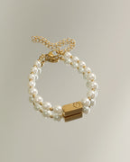 Pulsera de dos hileras de perlas blancas con adorno rectangular dorado y cadena de extensión, sobre fondo claro.