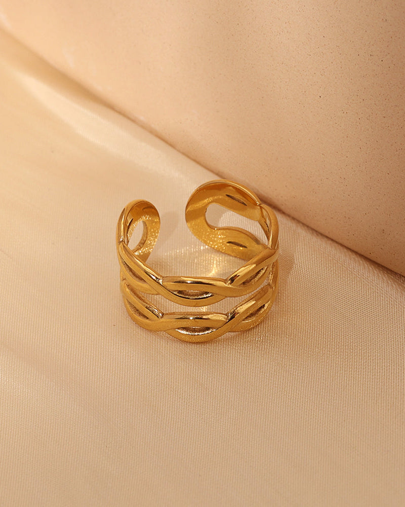 Anillo abierto ancho dorado con bandas onduladas entrecruzadas y huecos alargados, sobre tela beige.