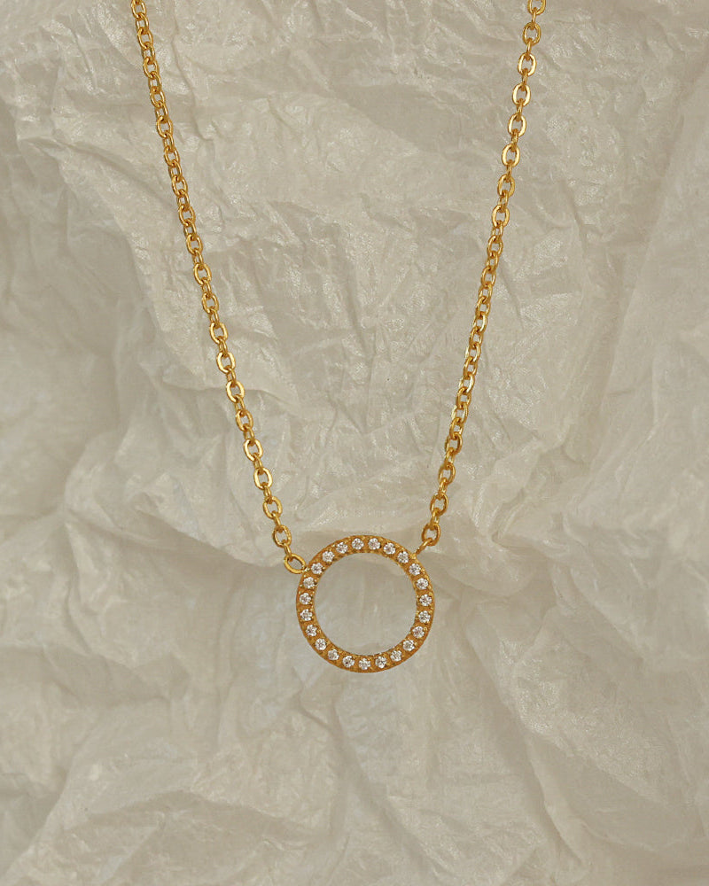 Collar de cadena fina dorada y círculo abierto con pequeñas piedras claras, sobre papel blanco arrugado.