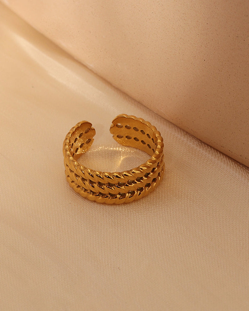 Anillo abierto dorado de varias hileras con textura entrelazada, sobre tela beige.