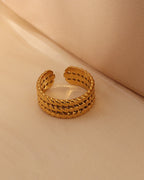 Anillo abierto dorado de varias hileras con textura entrelazada, sobre tela beige.