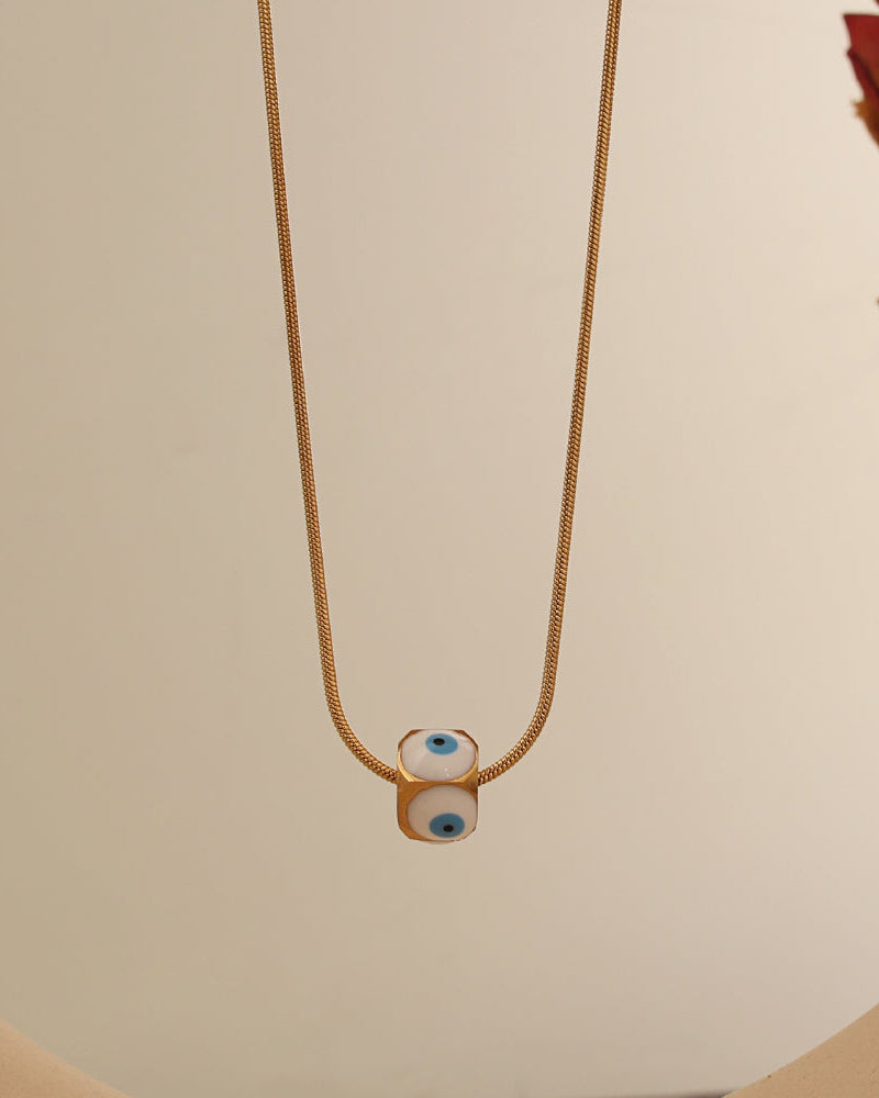 Collar dorado con dos pequeñas cuentas claras de motivo de ojo azul, suspendido sobre fondo crema.