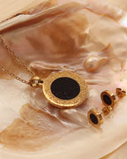 Collar y pendientes dorados de centro negro y contorno circular grabado, sobre una concha de tonos crema.