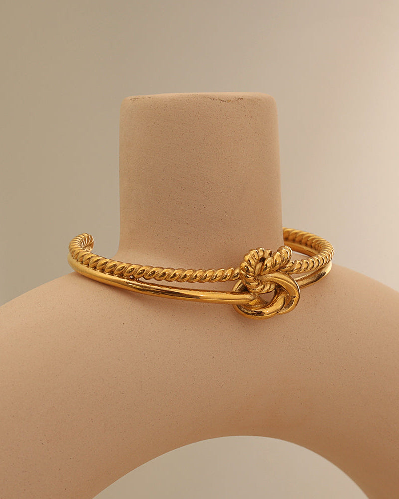 Pulsera rígida dorada de dos bandas con textura de cuerda y nudo central, sobre soporte beige.