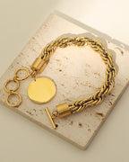 Pulsera dorada de cadena trenzada con disco redondo y cierre de barra, sobre una losa de piedra clara.