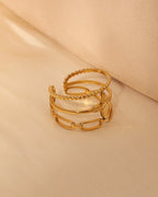 Anillo abierto dorado de varias bandas con texturas, motivos circulares y formas geométricas, sobre tela beige.