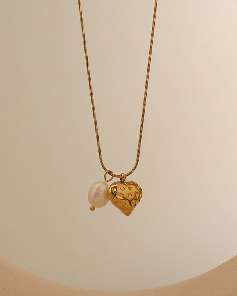 Collar fino dorado con colgante de corazón con textura y perla blanca, sobre fondo claro.