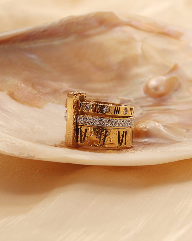 Anillo ancho dorado con números romanos y varias bandas de pequeñas piedras claras, sobre una concha.