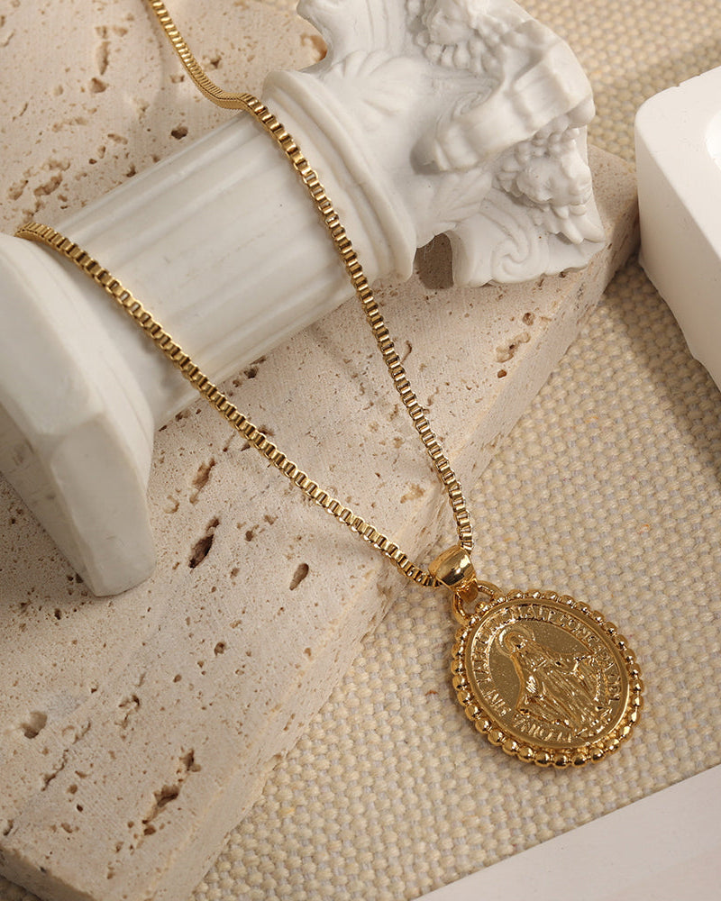 Collar de cadena dorada y medallón ovalado en relieve, sobre piedra beige junto a una columna decorativa blanca.