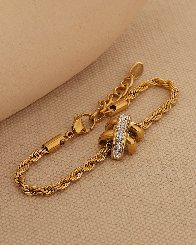 Pulsera dorada de cadena torcida con lazos y arco de piedras claras, colocada sobre tela beige.