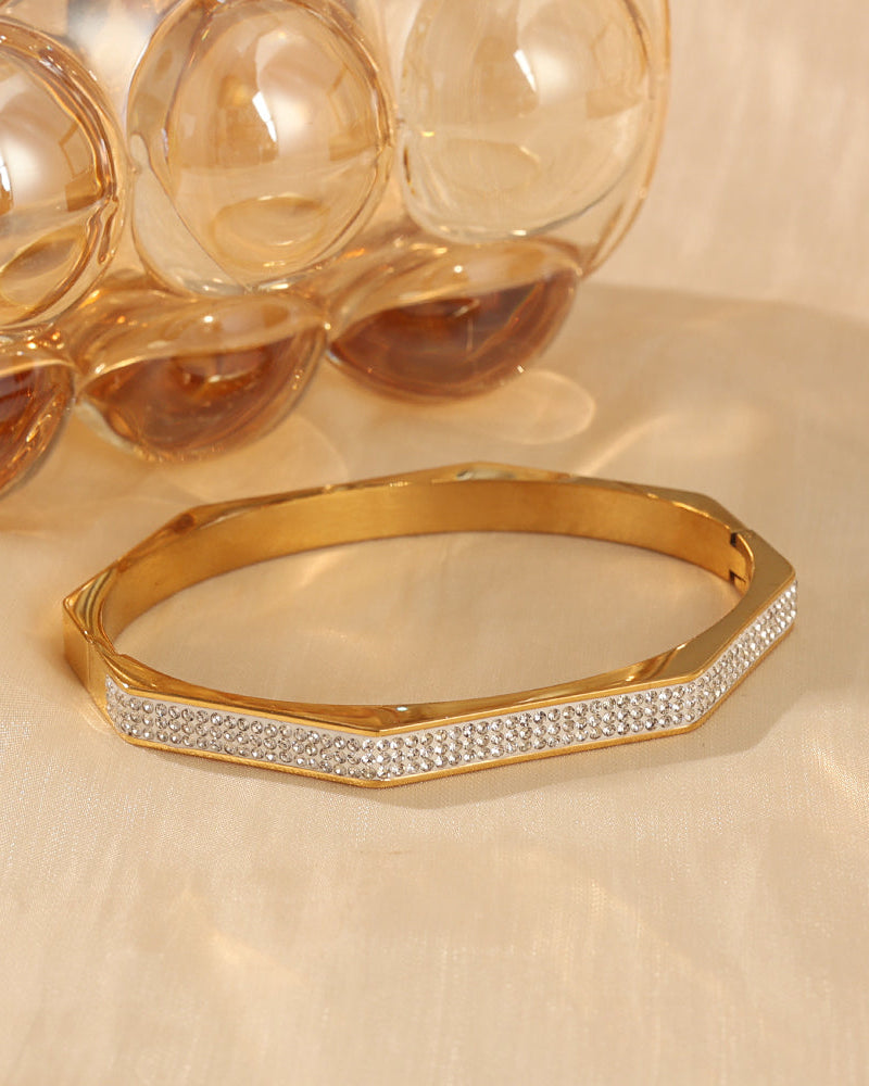 Pulsera rígida dorada de contorno anguloso y frente de pequeñas piedras claras, junto a recipientes de vidrio.